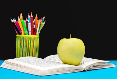 Kara tahtaya karşı renkli kalemlerle açık bir kitabın üzerinde yeşil elma. Okula geri dön..