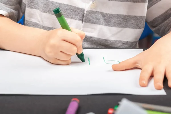 Drawing on a white sheet of paper with colored wax pencils. The concept of childrens creativity and hobbies. A child draws a green pencil on white paper. Baby hands closeup