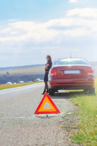 A red triangle an emergency stop sign against a blurred woman, the driver stands near her car and waits for help on the highway on a warm summer day. — Stock Photo, Image