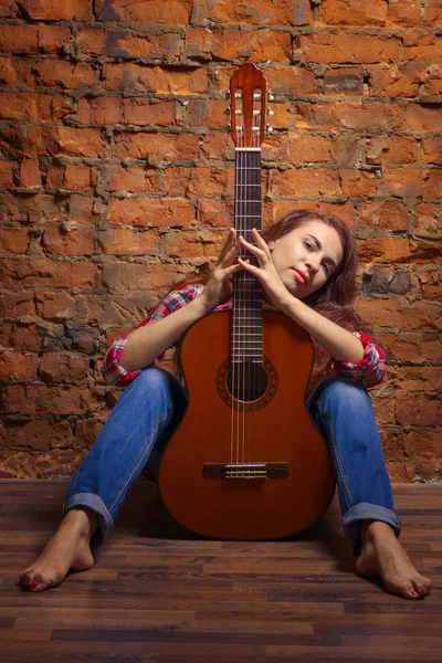 Uma jovem com os pés descalços senta-se contra uma parede de tijolos e abraça uma guitarra. Concerto de liberdade criativa — Fotografia de Stock