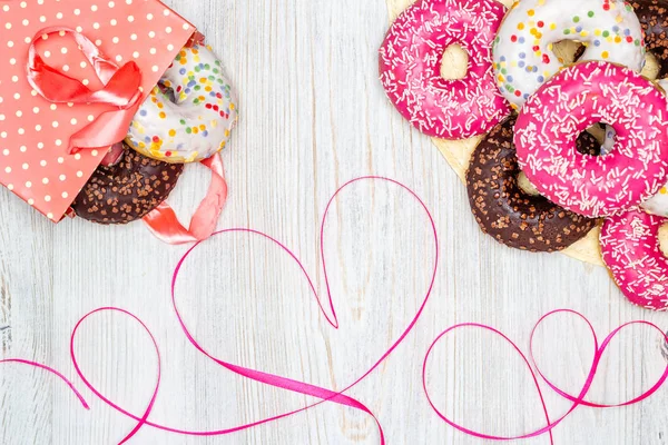 Donuts, gift bag with donuts inside and a red ribbon heart on a wooden table. Flat lay. Valentine\'s Day celebration concept.