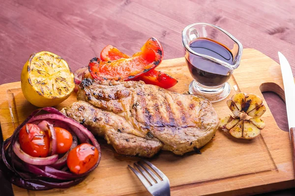 Carne de cerdo jugosa con una ramita de tomillo fresco, limón y verduras a la parrilla y salsa servida en una tabla de madera — Foto de Stock