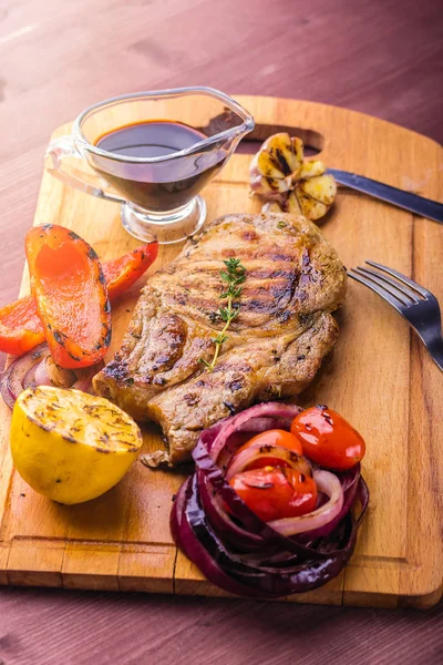 Carne de cerdo jugosa con una ramita de tomillo fresco, limón y verduras a la parrilla y salsa servida en una tabla de madera — Foto de Stock
