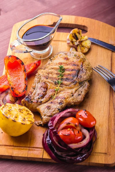 Carne de cerdo jugosa con una ramita de tomillo fresco, limón y verduras a la parrilla y salsa servida en una tabla de madera — Foto de Stock