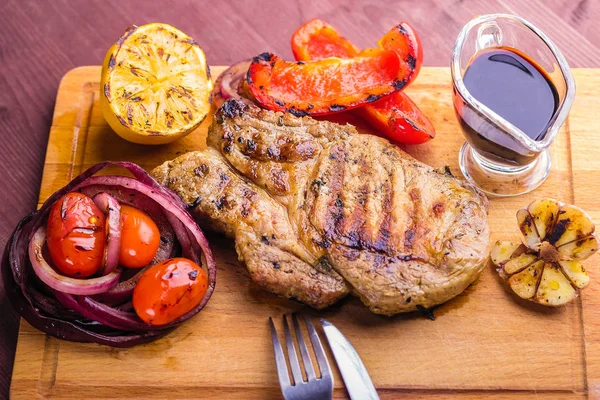 Carne de cerdo jugosa con una ramita de tomillo fresco, limón y verduras a la parrilla y salsa servida en una tabla de madera — Foto de Stock