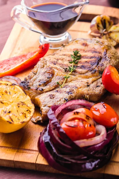 Carne de cerdo jugosa con una ramita de tomillo fresco, limón y verduras a la parrilla y salsa servida en una tabla de madera — Foto de Stock