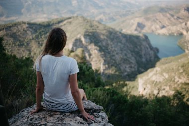 Genç ve güzel kız beyaz T-shirt ve zümrüt bir göl ile taş güzel kanyon yukarıda İspanya'günbatımı üzerinde oturan gözlüklü beyaz şort. Gezgin dağlarda.