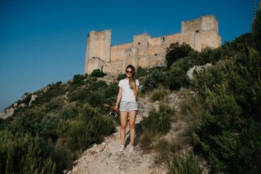 Sevimli genç kadın beyaz T-shirt ve beyaz şort dağlarda arka planda antik kale ile kumlu yollarda on sunset İspanya'da kalıyor. Gezgin dağlarda.