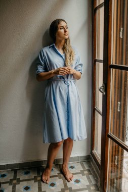 Düz zemin üzerinde beyaz duvarlı İspanya pencere bakarak poz uzun mavi elbiseli güzel esmer Bayan