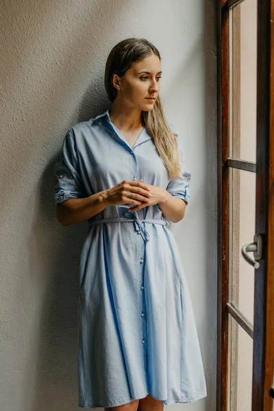 Sevimli genç kadın düz zemin üzerinde beyaz duvarlı İspanya pencere bakarak poz uzun mavi elbiseli 