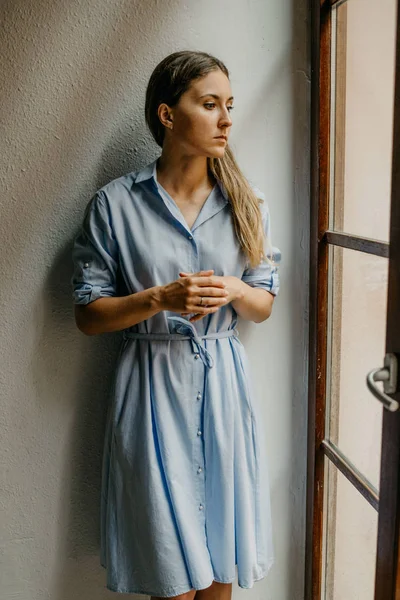 Şirin esmer kadın düz zemin üzerinde beyaz duvarlı İspanya pencere bakarak poz uzun mavi elbiseli