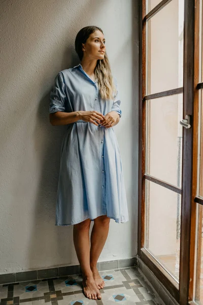 Düz zemin üzerinde beyaz duvarlı İspanya pencere bakarak poz uzun mavi elbiseli güzel genç kadın