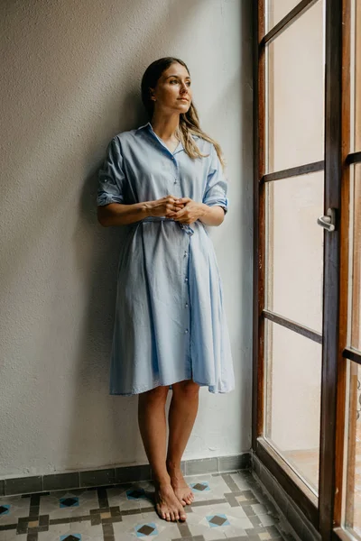 Uzun Mavi elbiseli Genç ve güzel kadınlar beyaz duvarlı İspanya pencere bakarak düz yerde duruyor