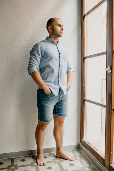 Tanned brutal man in the blue shirt and blue jeans shorts posing holding hands in pockets in the flat with white walls looking out the window in Spain