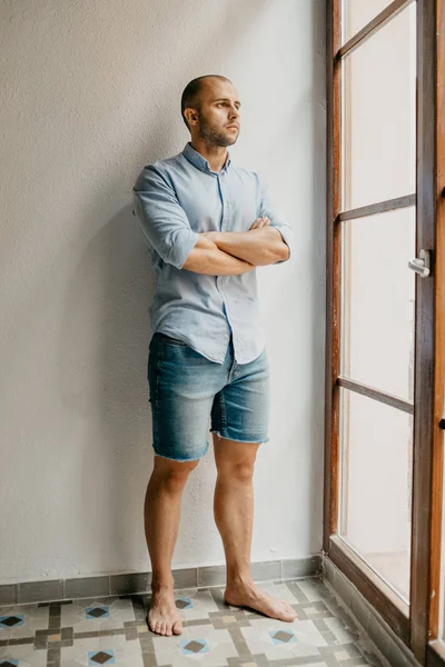 Tanned attractive man with arms crossed on the chest in the blue shirt and blue jeans shorts posing holding hands in pockets in the the flat with white walls looking out the window in Spain