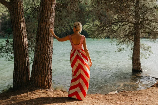 山の湖の近くの古い木の近くポーズ赤と白のドレスでスタイリッシュな魅力的な金髪の女性 後ろからの肖像画 スペイン — ストック写真