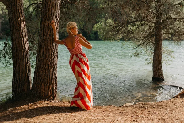 山の湖の近くの古い木の近くの赤と白のドレスでスタイリッシュな魅力的な金髪の女性 後ろからの肖像画 スペイン — ストック写真