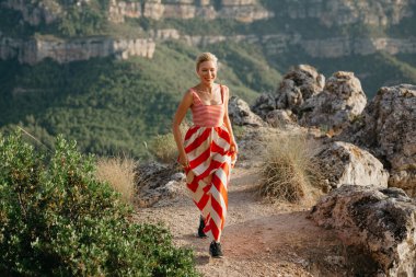 Bir güzellik sevimli sarışın Bayan uçurumun tepesinde Dağı arka planda çalışan kırmızı ve beyaz elbiseli. Mountain view. Akşam İspanya. Kanyonu Panoraması