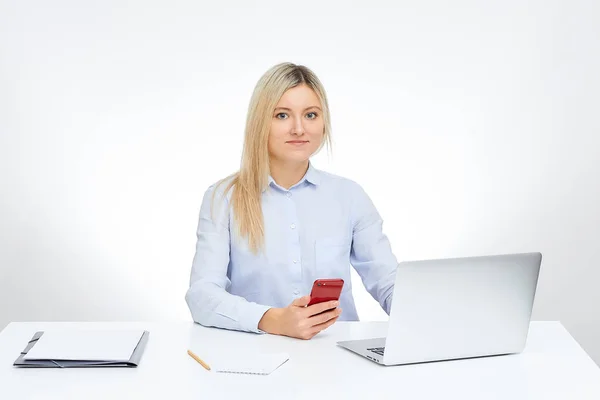 Young blonde woman watches straight holds her red glass cell phone works with her aluminum notebook computer and sits by the desk in the office with the white background. Documents on the table.