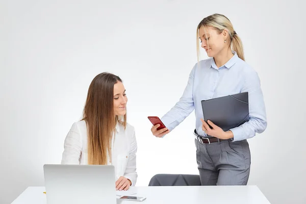 Blonde caucasian female manager shows something on the cell phone to her brunette female colleague with a smile in the office with the white background.