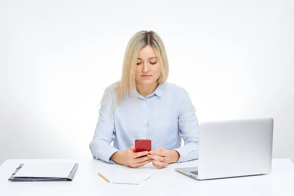 年轻的金发女子用红色玻璃手机阅读新闻 坐在办公室的办公桌旁 背景是白色的 台式机和桌子上的文件 — 图库照片