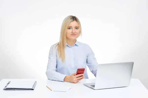 年轻的金发碧眼的女人直视着她的红色玻璃手机与她的铝笔记本电脑一起工作 坐在办公室的办公桌旁 背景是白色的 表中的文档 — 图库照片