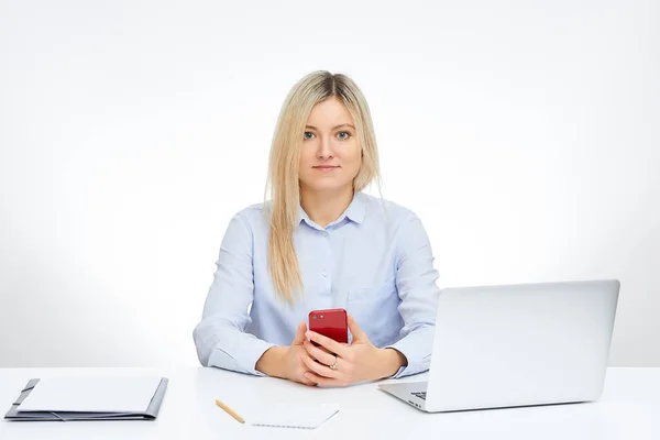 Young blonde woman watches straight holds her red glass cell phone and sits by the desk in the office with the white background. Aluminum notebook computer and documents on the table.