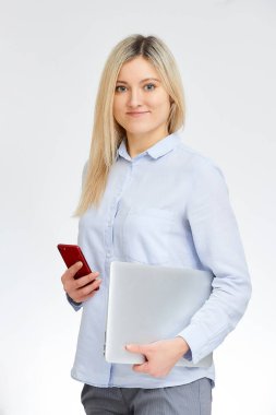 Sevimli sarışın genç beyaz kadın gülümseme standları yan tek kollu alüminyum dizüstü bilgisayarını tutar ve başka bir beyaz bir arka plan ile ofiste kırmızı cam cep telefonu tutar.