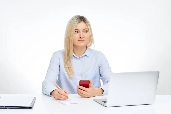 年轻的金发碧眼的女人看着拿着她的红色玻璃手机 拿着一支铅笔 坐在办公室的桌子旁 背景是白色的 台式机和桌子上的文件 — 图库照片