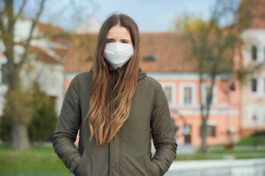 Uzun saçlı bir kadın, koronavirüsün yayılmasını önlemek için tıbbi yüz maskesi takar. Yüzünde ameliyat maskesi olan bir kız eski bir kasabada COVID-19 pozu veriyor..