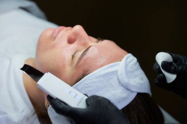 Kozmetoloji ofisinde sıcak bir losyondan sonra kadının yüzünün yakın plan fotoğrafı. Tek kullanımlık tıbbi eldivenlerle kozmetolojistin elleri ultrasonik deri fırçası ve pamuk tutuyordu..