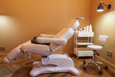 Kozmetoloji ofisinin modern iç mekanı. Tam elektrikli yüz güzelliği yatağı ve sandalyesi var. Salon taburesi, cilt bakım ürünleriyle dolu bir tramvay arabası ve güzellik salonunda bir lamba..