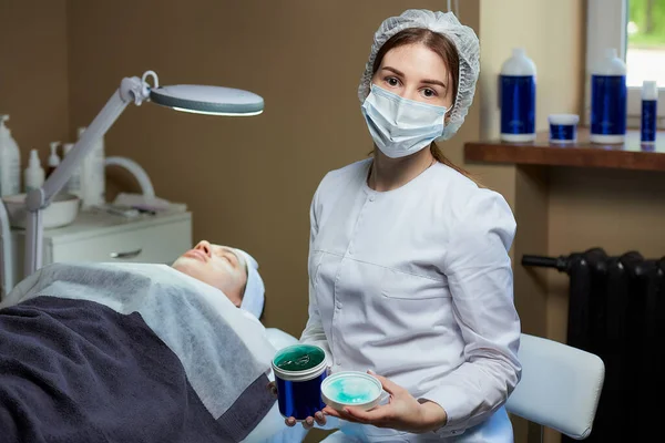 A female cosmetologist in a surgical face mask posing with a skincare gel near a patient on a full-electrical beauty bed in a cosmetology office. A procedure for skin cleaning in a beauty salon.