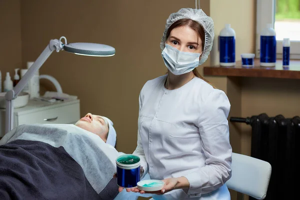 Tıbbi maske takan kadın bir kozmetik uzmanı kozmetoloji ofisinde elektrikli bir güzellik yatağında bir hastanın yanında deri jeliyle poz veriyor. Güzellik salonunda deri temizleme prosedürü..