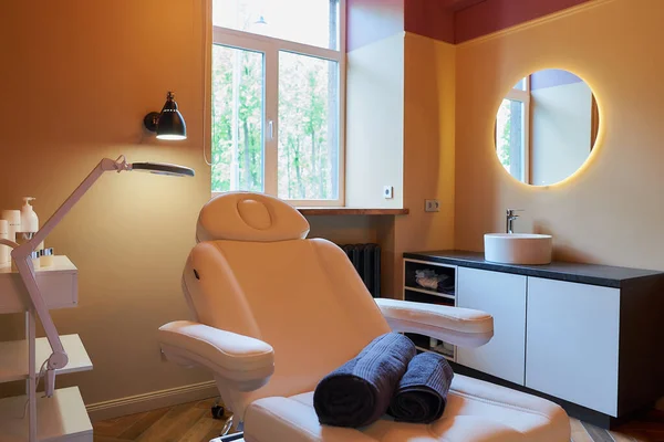 Interior of the cosmetology office with a sink, a round mirror, an electrical facial beauty bed and chair, a trolley cart with skincare products, a led lamp in a beauty salon. A window curtain raised.