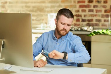 Sakallı bir adam evde kol saatine bakıp kredi kartı tutuyor. İnternette ödeme yapmak için bekleyen bir adam.. 