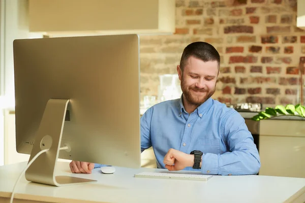 A happy man with a beard looking at his wristwatch during an online video briefing with his colleagues at home. A guy with a smile working remotely on a desktop computer in a private apartment.