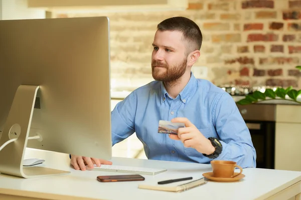 Hombre Con Barba Sienta Frente Computadora Sostiene Una Tarjeta Crédito —  Fotos de Stock