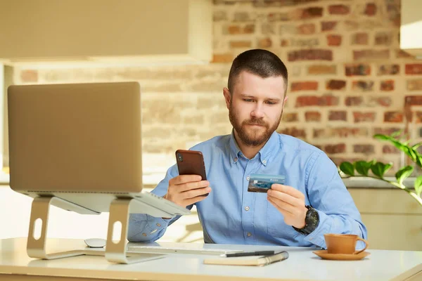 一个留着胡子的男人从信用卡后面看信息 然后在智能手机上输入 然后在家里上网购物 一个在公寓里用手机上网付款的人 — 图库照片