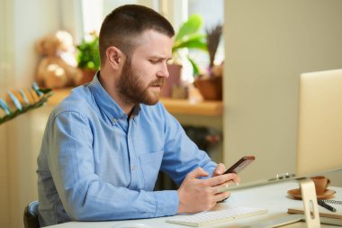 Mavi gömlekli somurtkan bir adam internette dizüstü bilgisayarın önünde akıllı telefondan haber arıyor. Evden uzakta çalışan sakallı bir adam.. 