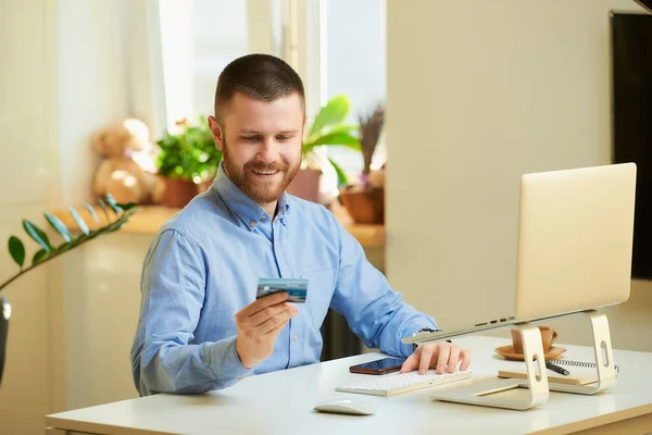 A happy man smiling and looking at his credit card in front of the laptop computer at home. A guy in a blue shirt with a beard doing an online payment on the internet on a laptop in his apartment.