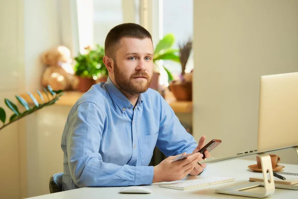 A puzzled man sitting at a white desk in front of a laptop and holds a smartphone and a credit card in his hands at home. A guy doing an online payment on the internet on a laptop in his apartment.