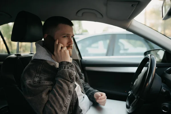 A man with a beard doing business calls on his smartphone inside a comfort car, a laptop computer lies on his lap. A guy stopped his car to immediately remotely solve tasks at work in social distance.