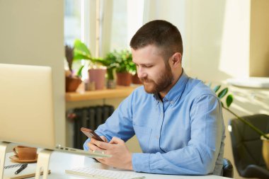 Mavi tişörtlü bir adam dizüstü bilgisayarın önünde akıllı telefondan haber arıyor. Evden uzakta çalışan sakallı bir adam.. 