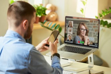 Bir erkek çalışanın, iş arkadaşlarını uzaktan dinlerken dizüstü bilgisayarla yaptığı bir görüşmenin ardından evde akıllı bir telefonla iş yaparken görüntüsü. Çevrimiçi bir toplantıda bir iş ekibi.