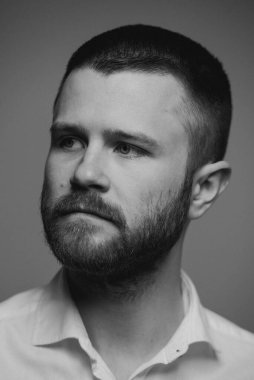 Gri bir arka planın önünde duran beyaz gömlekli, sakallı, vahşi bir adamın yakın portresi. Ciddi bir adam başka tarafa bakıyor. Siyah beyaz bir fotoğraf..