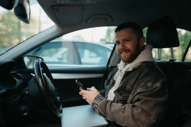 Arabada akıllı telefonuyla iş yapan sakallı bir adam dizüstü bilgisayarı kucağında yatıyor. Gülümseyen bir adam iş yerindeki görevleri uzaktan çözmek için arabasını durdurdu.