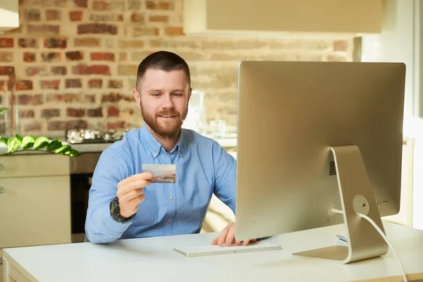 A man with a beard types a credit card number to do online shopping at home. A guy doing an online payment on the internet on a desktop computer in his apartment.