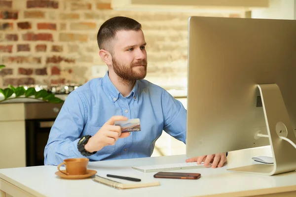 A man with a beard sits in front of the computer and holding a credit card at his hand at home. A guy doing an online payment on the internet on a desktop computer in his apartment.