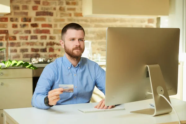 A man with a beard sits in front of the computer and types a credit card information on an online store at home. A guy doing an online payment on the internet on a desktop computer in his apartment.